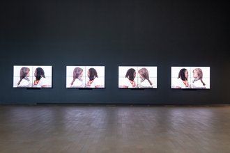 Installation view of four screens aligned on a wall. Each screen shows Stephanie Dinkins facing Bina48 on a white background