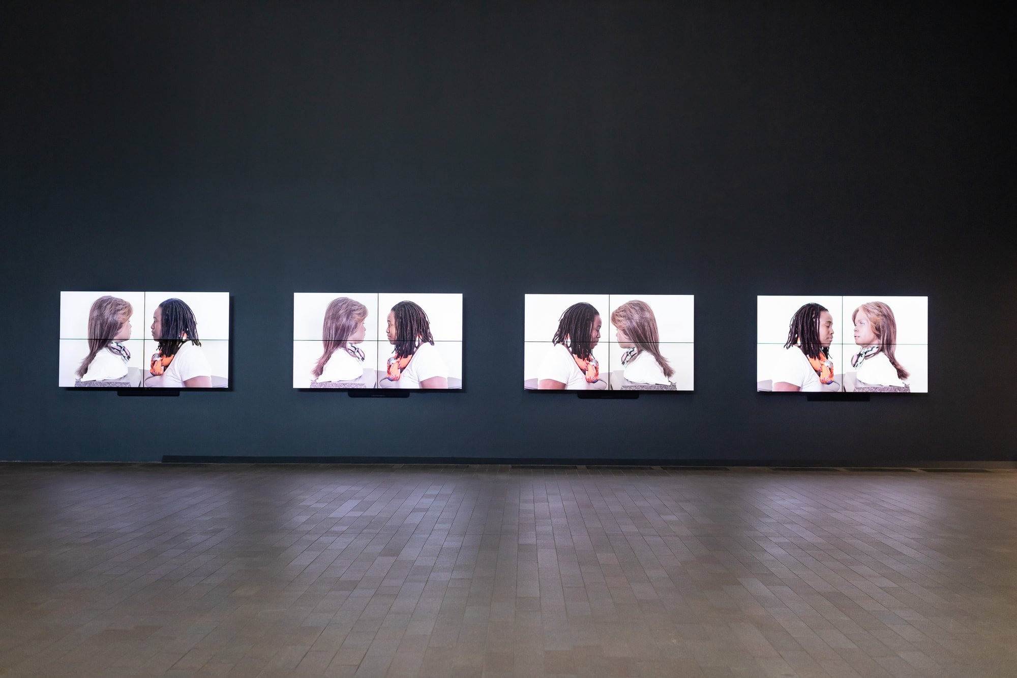 Installation view of four screens aligned on a wall. Each screen shows Stephanie Dinkins facing Bina48 on a white background