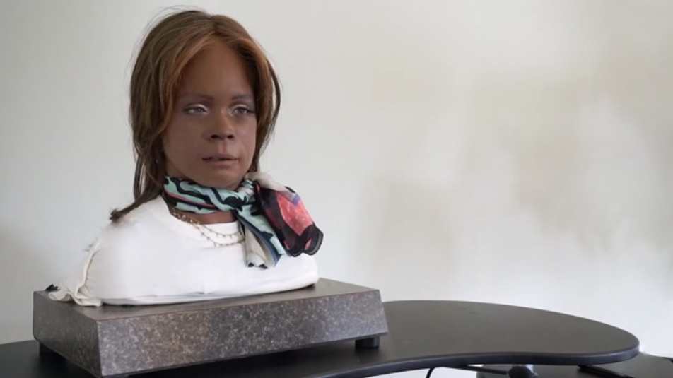 A bust of Bina 48 sitting on a table. Bina48 has dirty blonde hair, dark skin, a white t-shirt, and a scarf tied around her neck