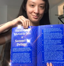 Victoria Sin smirking into a camera while holding the open book to a blue spread that shows an interview with Samuel Delany