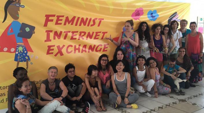 Photo of a group of people smiling for the camera in front of an orange "Feminist Internet Exchange" banner that has a graphic illustration of a woman holding a tablet and wearing a red cape.