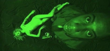 An aerial shot green filtered photo with a naked woman lying seductively a fabric covered floor and a black man wearing a tie and a dress shirt looking up to the camera.