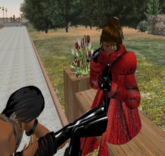 Two avatars in Second Life. One mistress is clad in a laytex bodysuit and a red coat. A submissive is kneeling at her feet, massaging her leg.