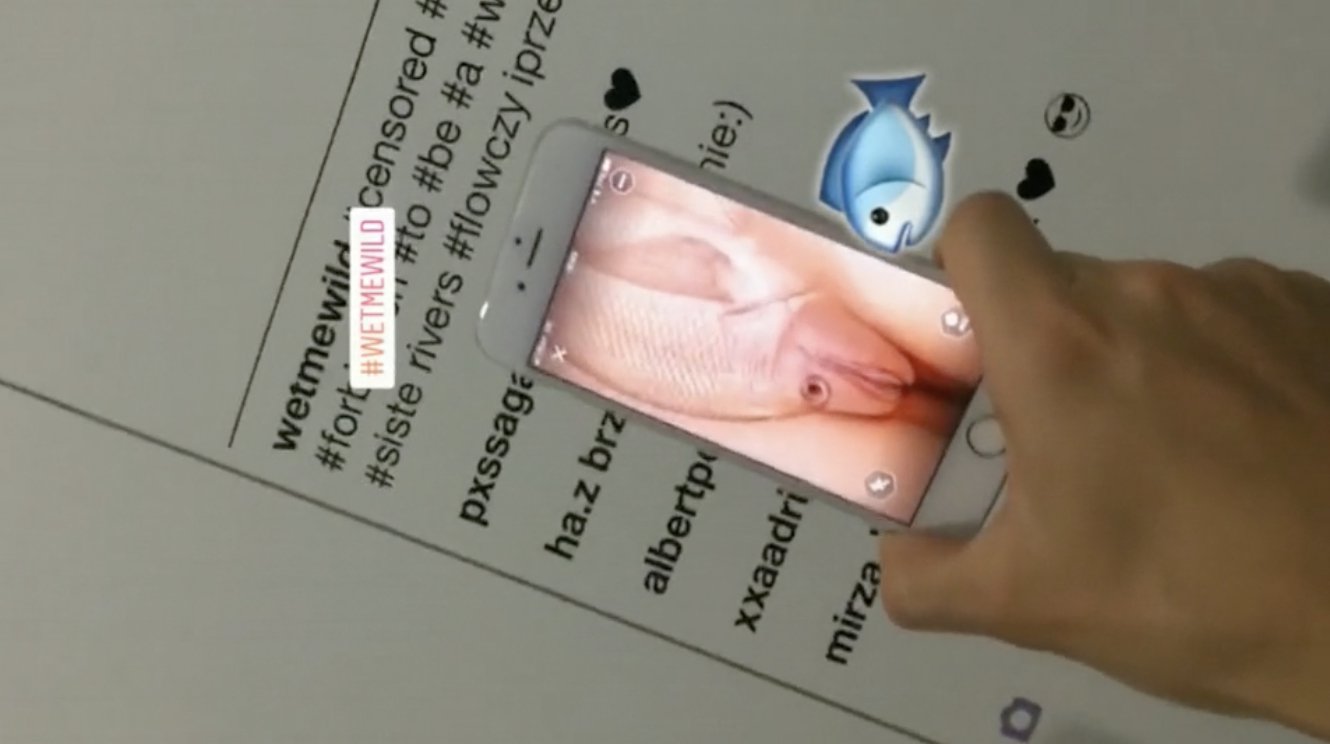 a hand holds a phone above wall text. The phone displays what appears to be a vulva that is actually a light pink fish