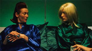 Two people elegantly lays back on green silk pillows in a room with green walls. The person on the left with half black and white hair wears royal blue silk, hands crossed, talks to the person on the left with long blonde hair wearing a green silk.
