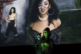 A light-skinned woman with curly hair under a green spotlight stands in front of a backdrop of two of her portraits