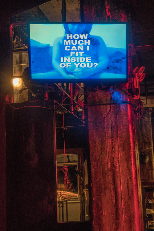 an installation of a blue screen with a woman sitting beneath white text. The screen is high in an industrial setting