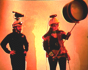 Two people in an orange-lit room. One with a mustache, hands on hips, and a headlamp and video camera attached to the helmet observes the person wearing sunglasses to the right, who has a projector on their head, carrying a cylindrical screen by a stick.