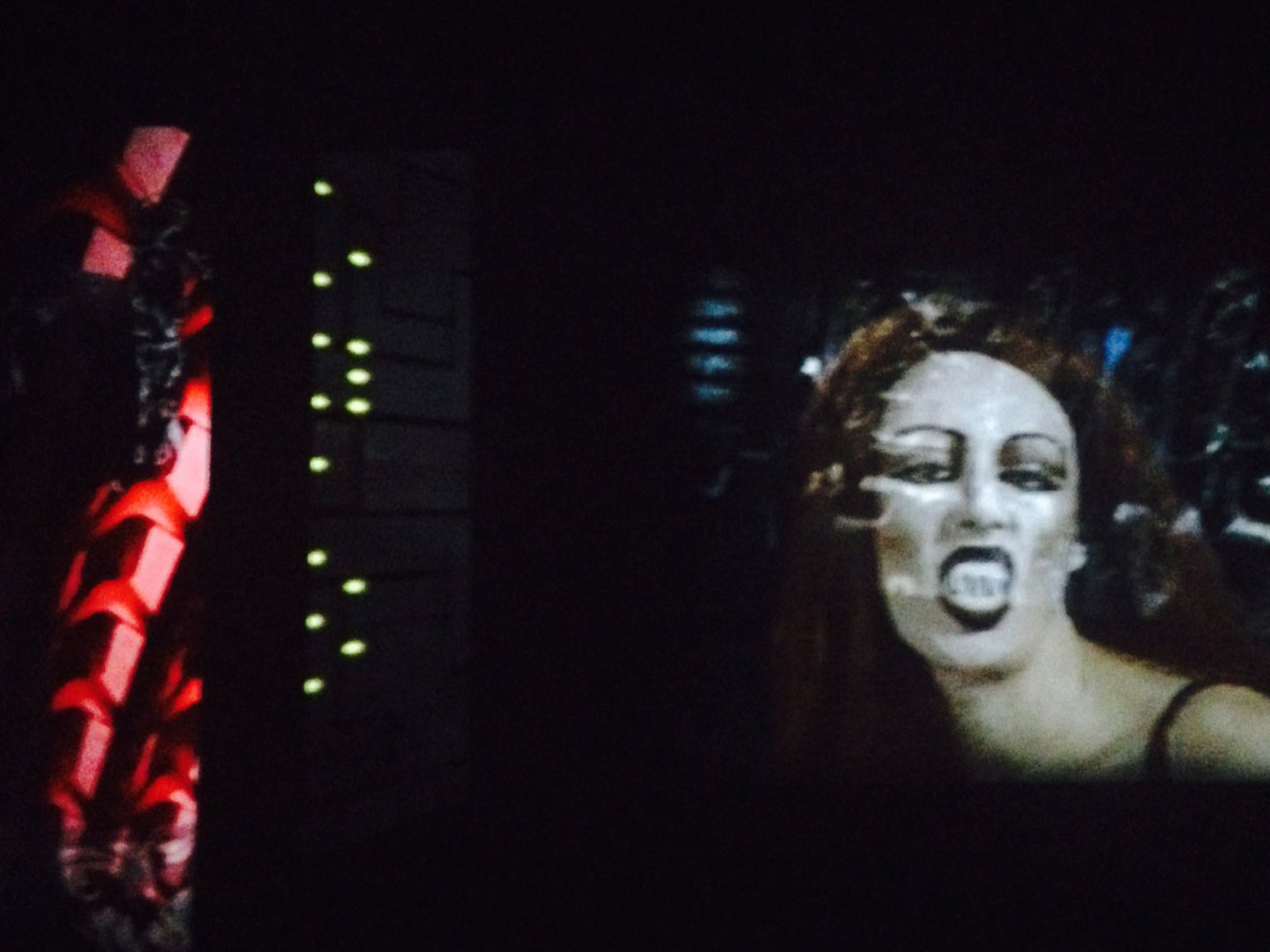 A photo collage of a woman with long brown hair and dark lipstick opening her mouth layered onto a dark black room with blue and yellow lights twinkling from a machine. A red bionic tail like figure flanks the left.