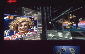 An image of a cyberpunk virtual world showing a screen of a news anchor woman in a blonde curled bobbed wig smiling with "Today 10am" typed in white below on the left and a screen of two women kneeling in the midst of a dance on the right.