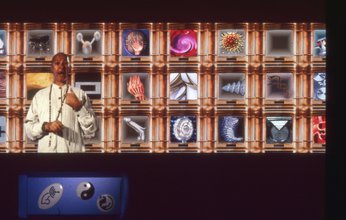 An image of a virtual world showing a man wearing white and a long beaded necklace pointing to himself as he speaks. Behind is a golden grid displaying different icons like a metallic dildo, a pink whirlpool, a globular virus and so on, inside the frame.