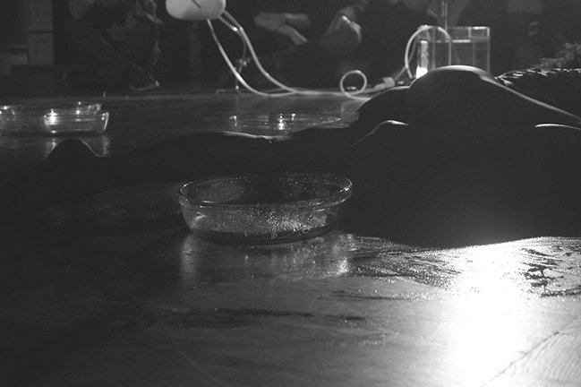 An eerie black and white photo of a naked body lying on the floor with empty plastic water bins. A machine connected to white tubes trailing to a filled water container sits in front of an sitting audience.