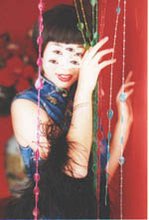 A photo of an Asian woman wearing a royal blue floral mandarin gown and a black feather boa on the arms, posing in a red room as she plays with colorful beaded curtains. The woman smiles and has five eyes, like that of a spider.