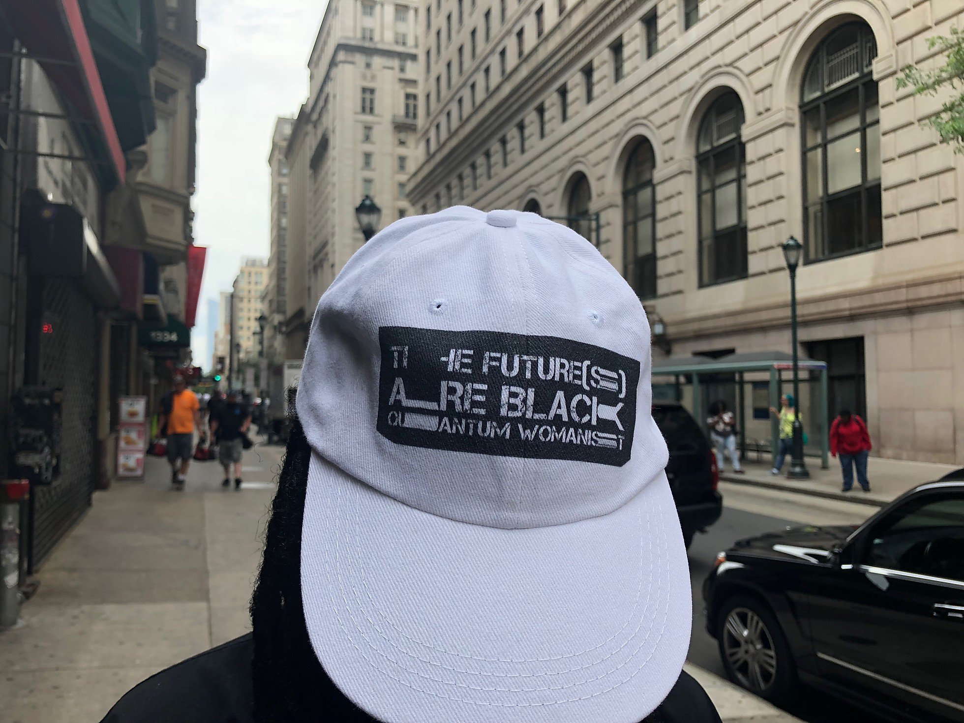 Photo of a person walking the streets of NYC wearing a white hat backwards, showing a black rectangle with "The future(s) are black quantum womanist" typed in distorted letters on the hat's front.