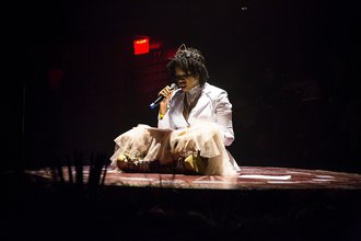 Photo of a performance of a woman with black curly hair sitting on the floor of a stage in a white blazer and a fluffy dress. She wears a crown on her head and speaks into a microphone while reading off of a paper on her lap.
