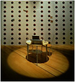 Photo of an installation with a large wall of red circles in columns and rows. A spotlight shines on a round table with an open briefcase assisted by two chairs.