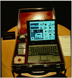 Photo of an open briefcase displayed on a wooden round table. Inside is a laptop largely in the right and a phone, a webcam, and a red button sectioned off into a column on the left of the briefcase.