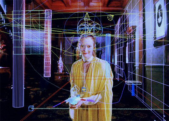 A woman wearing a yellow garment smiles into the camera while standing in the hallway to a large wealthy home. Computer drawings grids planes on the walls and columns of the building are scribbled on top.