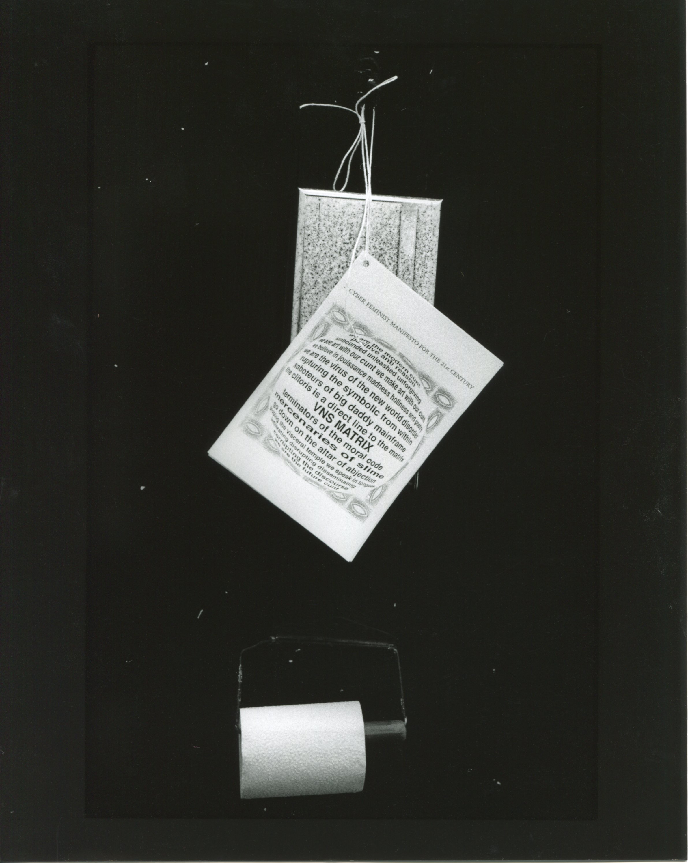 Black and white photo of the Cyberfeminist Manifesto printed on paper, hanging by a white string tied onto a black hook, right above a toilet roll.