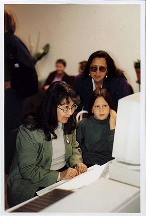 A photo of three women ranging in different ages, from elderly to middle aged to child, and races sitting around a computer and observing the screen.