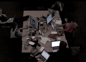 Photo of six people very focused and concentrated working on laptops or computers sitting around a large and cluttered table of coffee cups, chargers, wires, and snacks.