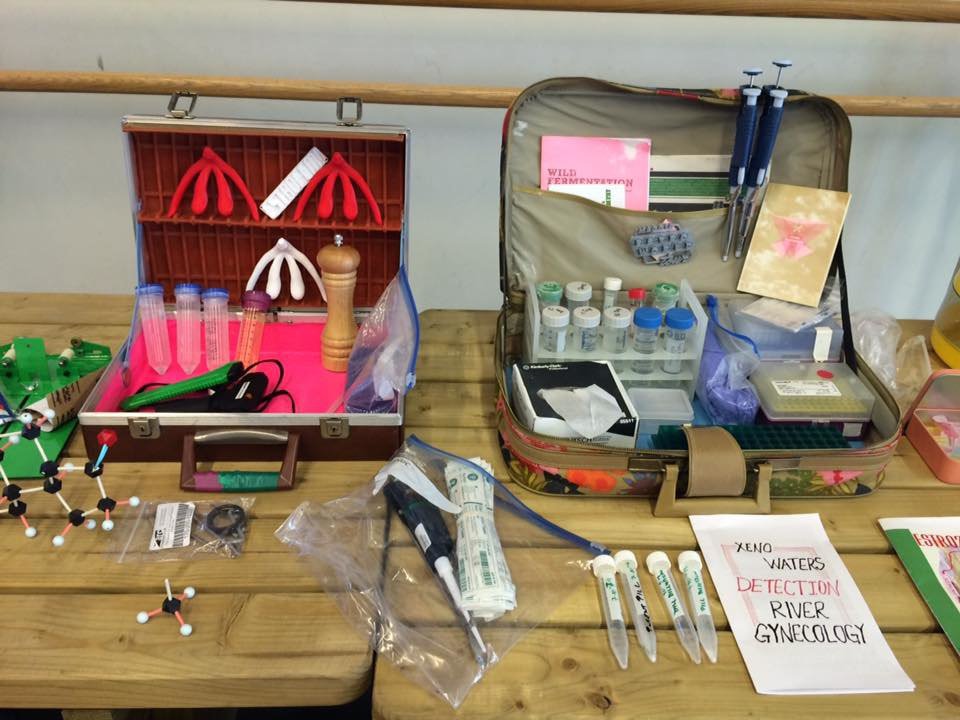 Two tables placed side by side with two briefcases opened, displaying lab equipment such as test tubes, syringes, molecular structures, booklets, and a pepper shaker.