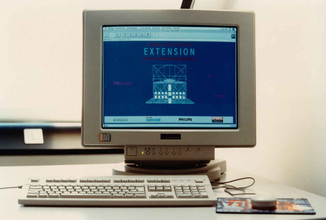 A photo of a retro desktop computer on a desk. The screen displays a blue filtered webpage with a red and white typed header and a gridded architectural geometric graphic largely in the center. Below is a row of conglomerate tech company logos.