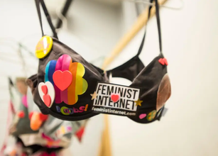 Photo of a black bra hanging on a rack covered in colorful and vibrant heart, flower, "boobs!" stickers with yellow circle and pink bow shaped pinned to its top corners. A black box sticker that says "Feminist Internet" is stuck to the middle of the bra.