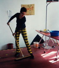 A candid photo of a person with short hair wearing glasses and yellow checkered pants sweeps the floor in an intimate shared space that has red floors, three sketches taped on the back wall, art materials scattered, and a surfboard shaped table.