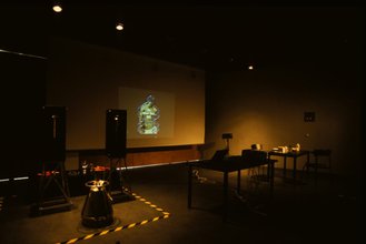 Photo of an installation with warm lighting, a projector screen of a small image of a male with DNA helixes wrapping his shirtless body, two desks on the right, and equipment bound by yellow and black striped tape on the floor on the left.