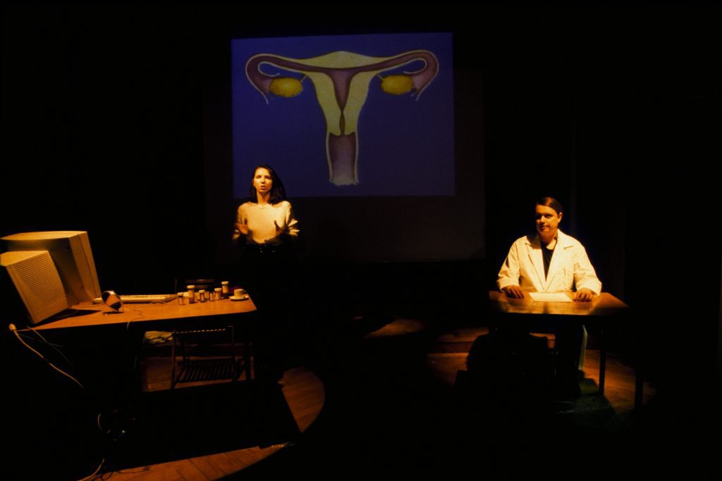 Photo of two people with long hair lit by warm theater lighting. One speaks with dramatic hand gestures next to a desk with a PC and pill bottles. The other wears a lab coat while sitting on a desk. A drawn uterus on a blue screen is projected behind.