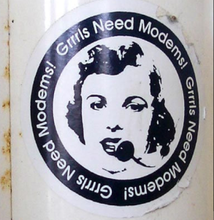 A black and white circle sticker. The text "Grrrls Needs Modems!" in white text and a black background borders a stenciled mid-century white woman talking into a headset.