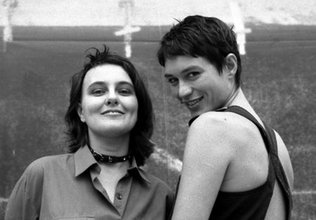 Black and white photo of two people smiling posing for a camera. The one to the left has long wavy hair and wears a collar choker. The one to the right has short hair and wears a black tank top.