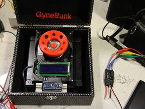 Photo of a black box surrounded by wires is labeled by a 3D printed red "GynePunk" sitting on the lid. A machine is placed on the bottom and has a large red wheel connecting to a blank screen screwed onto a circuit board. A smaller motherboard sits below.