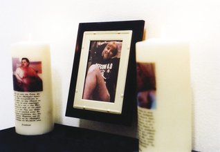 A framed photograph of a blonde woman with crossed legs wearing a black hoodie. The framed photo is flanked by two candles with a portrait of a man wearing a red shirt