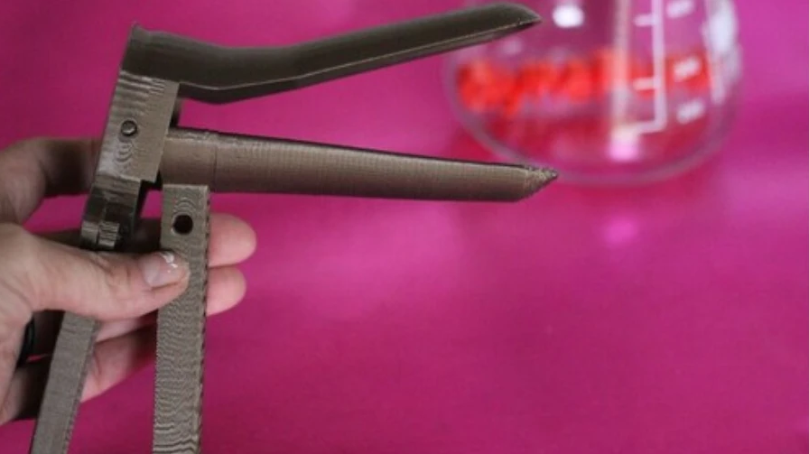 a hand holds a 3D printed speculum on a pink table