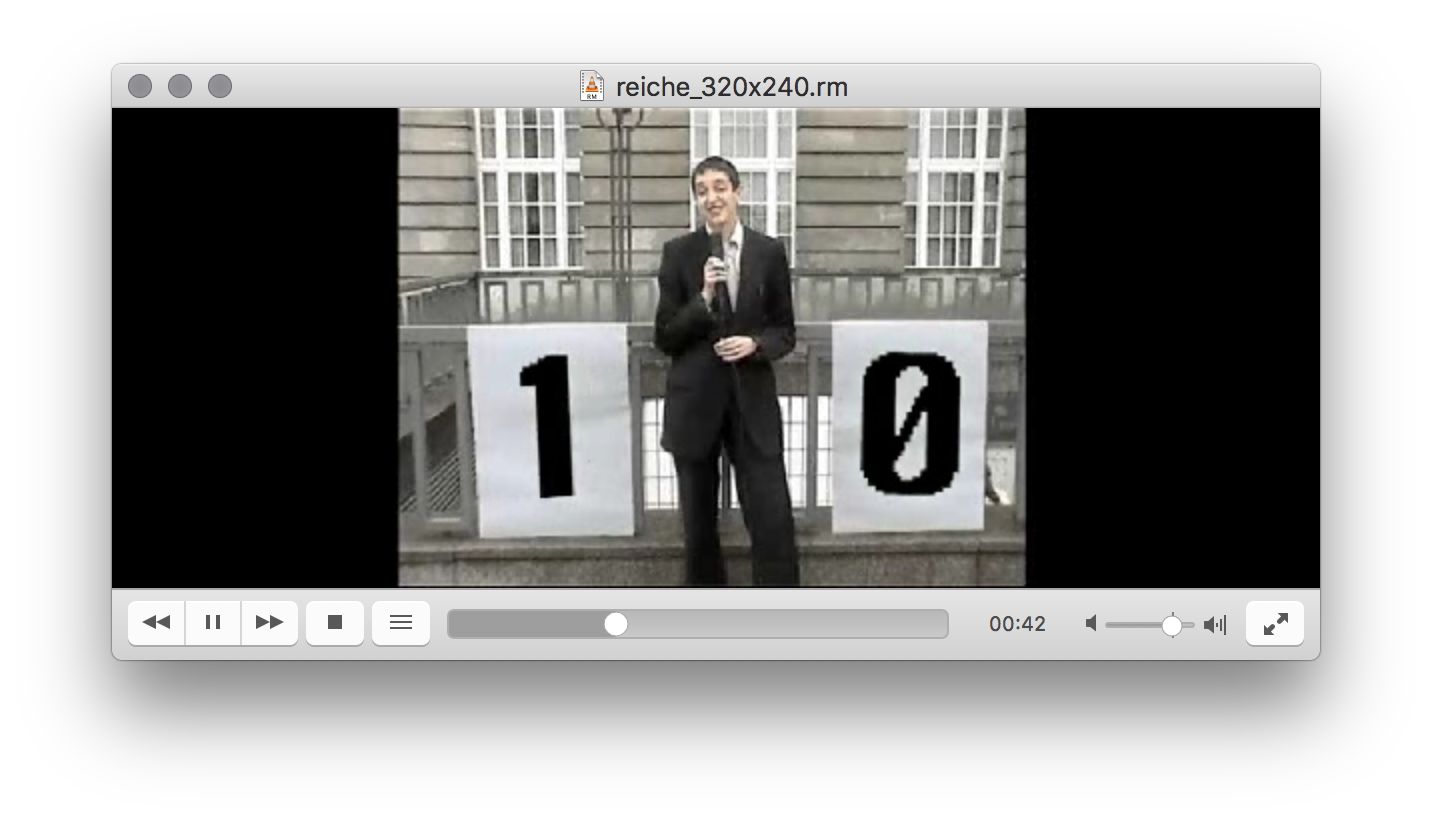 Screenshot of a video on a media player. A person wearing a suit smiles as she speaks into a microphone while standing in front of a grey building on an overcast day, in between the number 1 and 0 printed on white posters, taped to the fence behind.