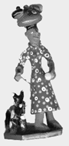 Black and white image of a table piece or sculpture of a cartoon figure wearing a polka-dot dress and balances a pile of clothes on her head. A shriveled dark animal crouches to the left.