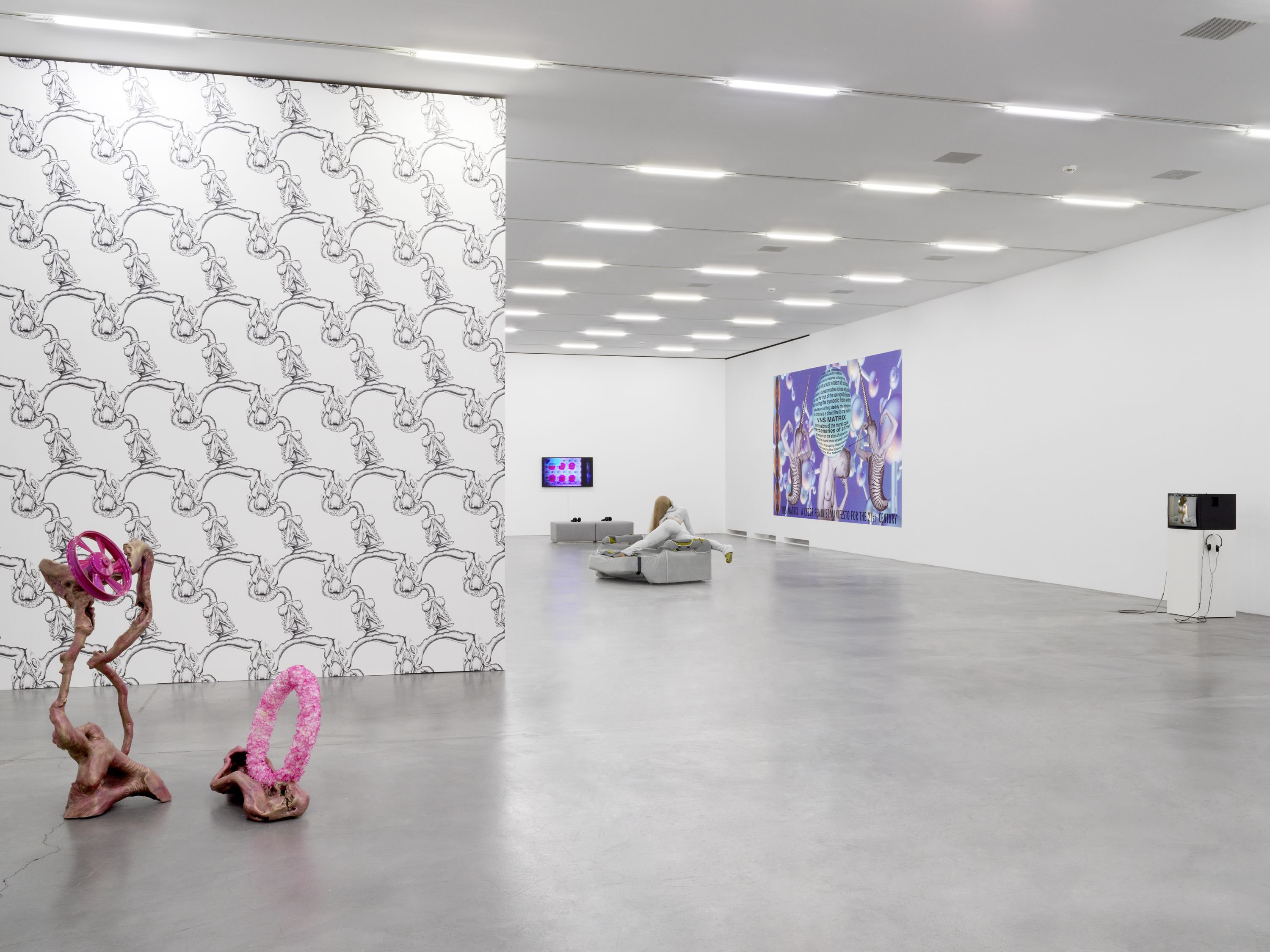 A bony sculpture holding a pink wheel and a ring sits in front of a large wall with skeletal patterns. On the right is a gallery large floor with TVs, a large VNS Matrix poster, and a mannequin wearing white lying twisted on a sofa, legs sprawled.