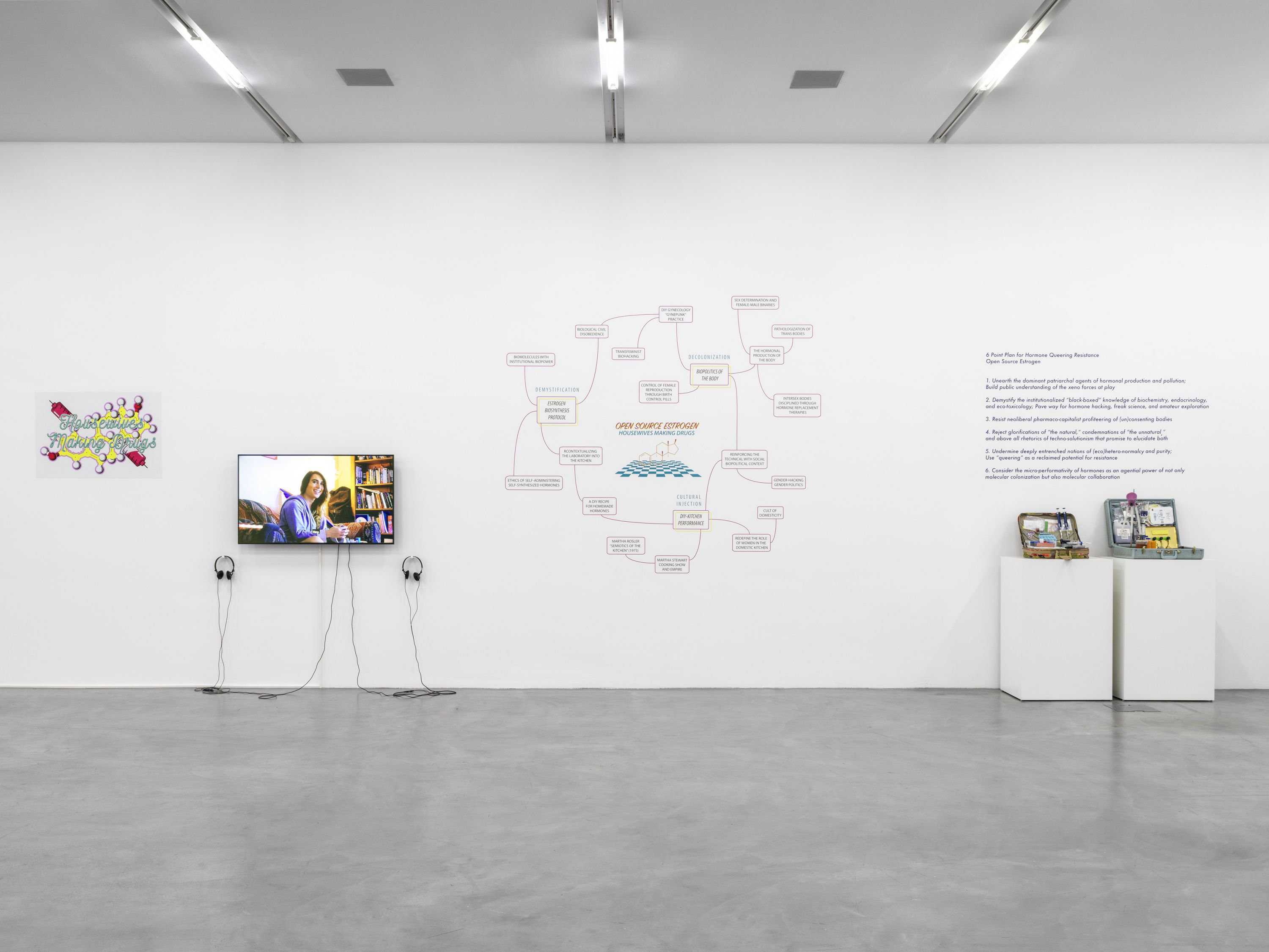 A gallery wall with a poster with molecules and red syringes, a TV, a poster of a flow chart, two white stands of open briefcases displaying lab equipment shown from left to right.