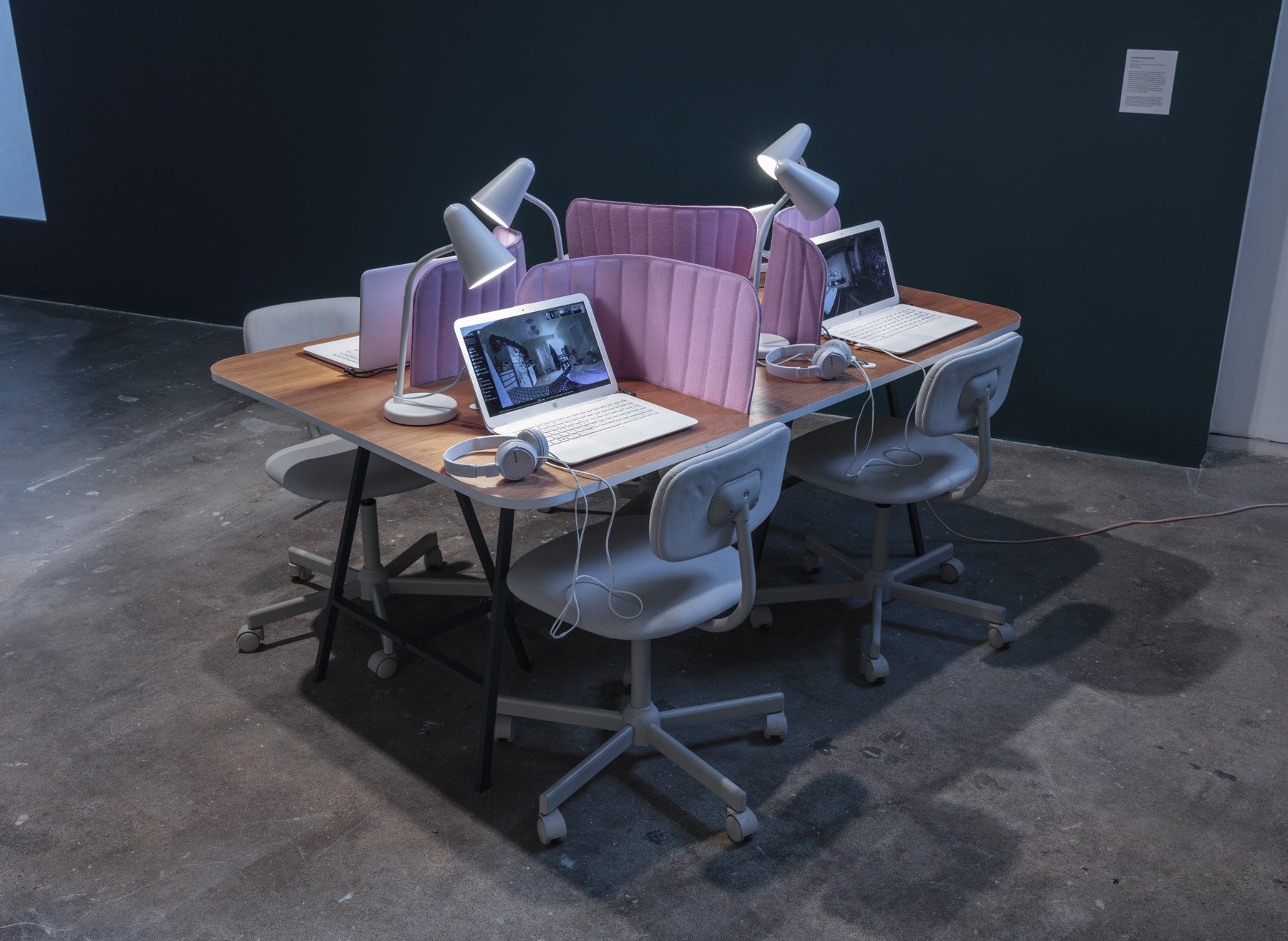An installation of a table with four seats that have identical white chairs, white open laptops with screens displaying virtual spaces, headphones, lamps, and pink foam desk dividers creating, creating designated and separate spaces per seat.