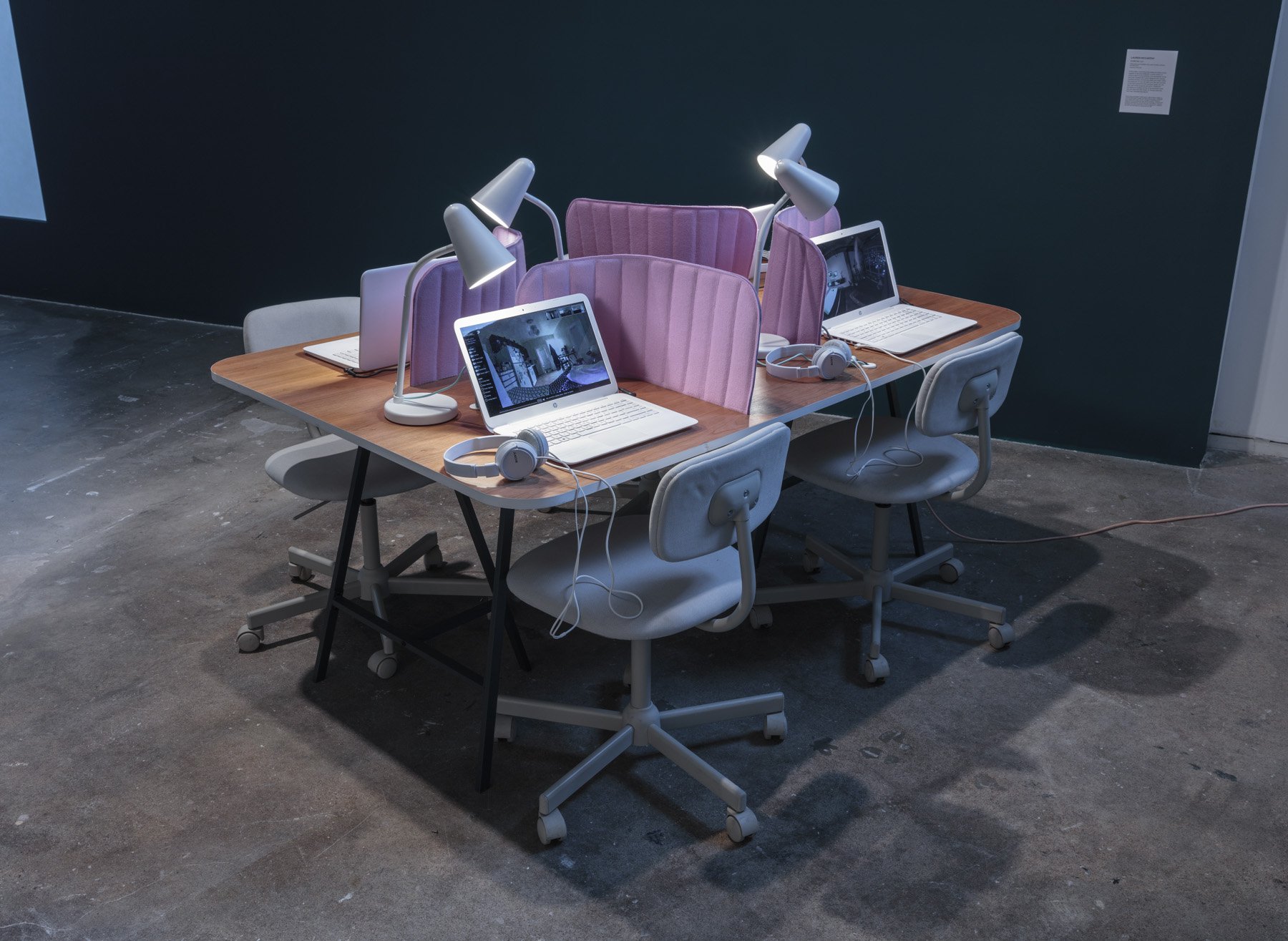 An installation of a table with four seats that have identical white chairs, white open laptops with screens displaying virtual spaces, headphones, lamps, and pink foam desk dividers creating, creating designated and separate spaces per seat.