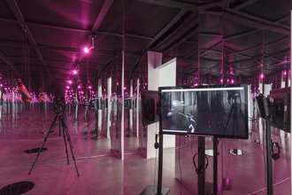 An installation of a mirrored wall, creating an infinitity room illusion. A pink light creates a pink filter. The right corner has a TV with headphones displaying a recording of what a camera beside it captures. A wooden object dangles from the ceiling.