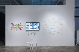 A large gallery wall with a TV screen displaying a show program of housewives making drugs. Two headphones and two chairs invite the viewer to watch. To the left is the logo enmeshed in molecules and syringes and a flow charts to the right.