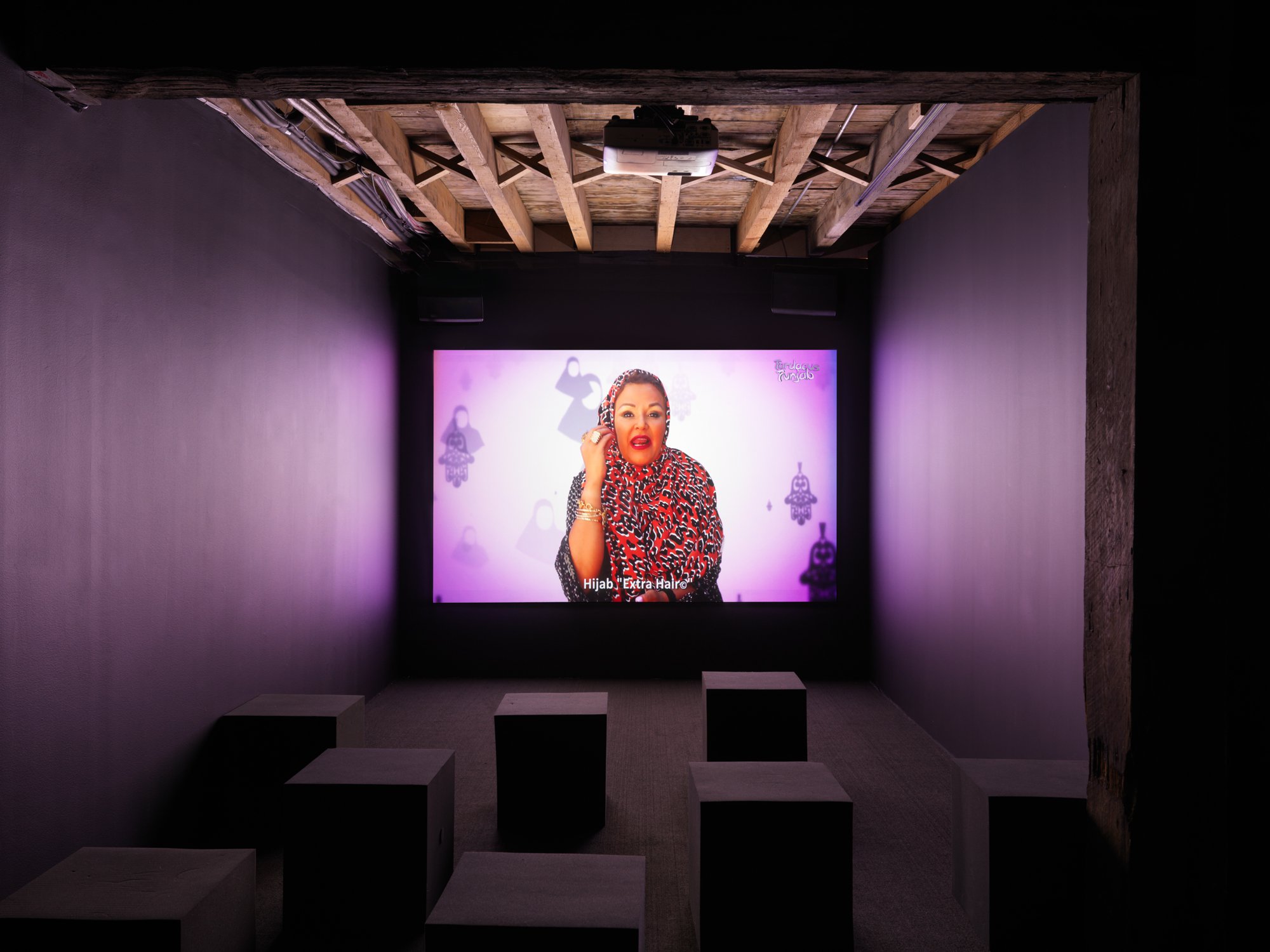 Photo of a small dark art gallery theater. Projected onto the wall is a  person wearing a red cheetah pint hijab talking on the phone in front of bright pink background with spiritual upside down Hamsa hands and silhouettes of a hijab.