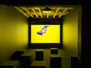 Photo of a small dark art gallery theater. Projected onto the wall shows zig zag cutout of legs in front of a yellow background, as if someone dove into a yellow screen.