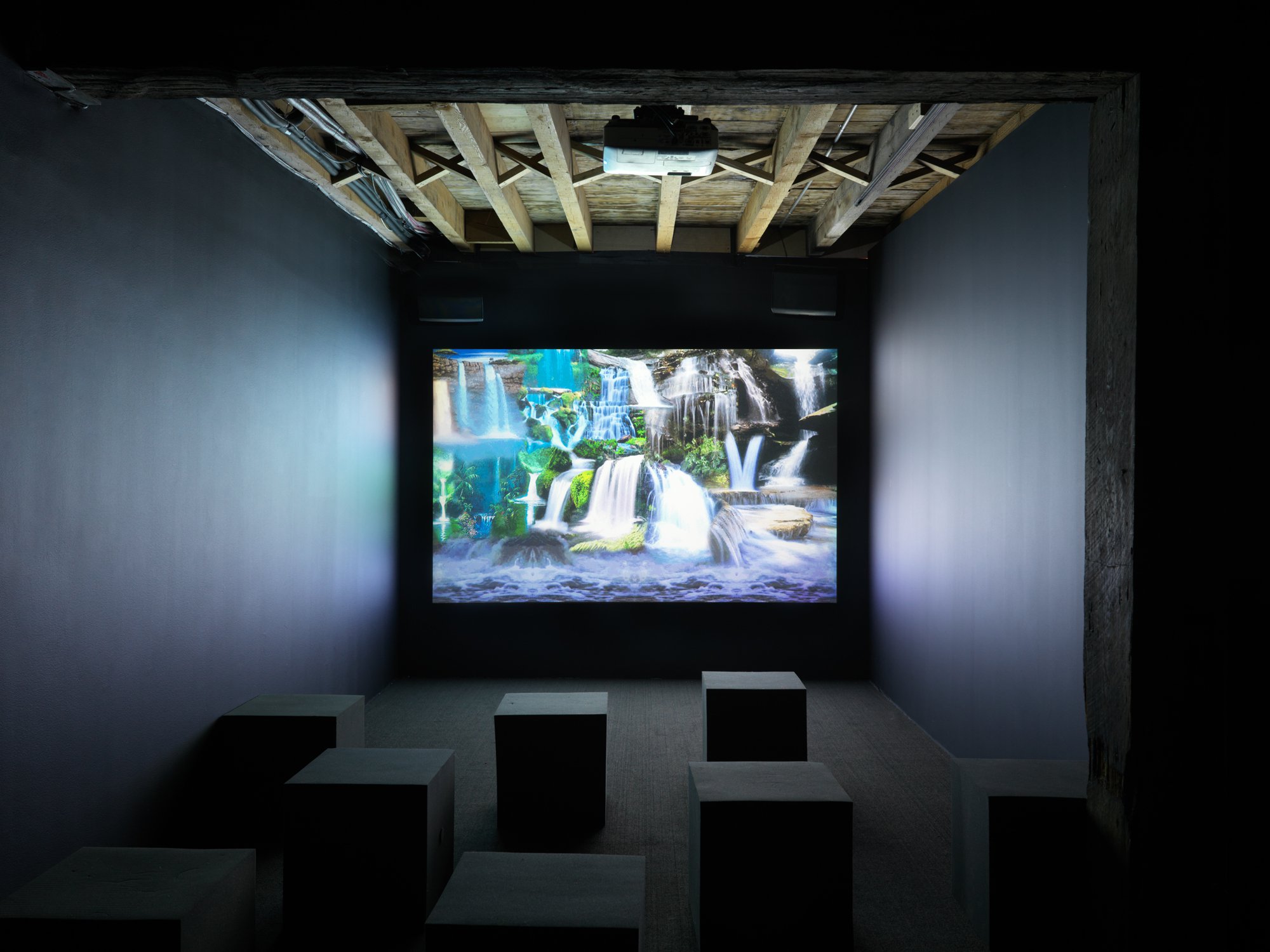 Photo of a small dark art gallery theater. Projected onto the wall is a scenic and serene view of many beautiful luscious waterfalls white and blue water flowing into a pool of water.