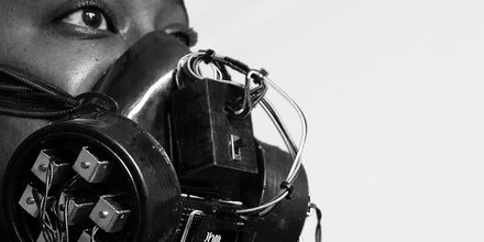 Black and white photogragh of person's face wearing a metallic clunky and industrial mask made of wires, covering nose and mouth.