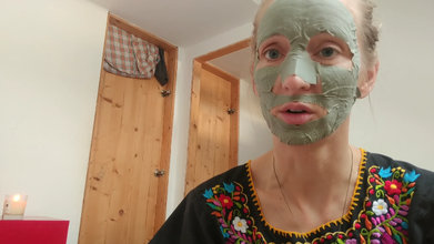 Image of a person with blond hair tied wearing a black top with colorful floral embroidery wears a clay skincare face sheet. They are in a room with a closed wooden door and a mirror behind them. A candle burns on a red desk to the left.