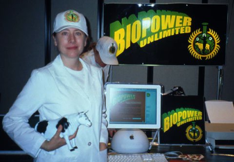 Photo of a woman wearing a white blazer, white top, and white hat while clutching a toy cow figurine poses. Behind her is a stand of Biopower unlimited posters and an old Mac PC with the Biopower site open.